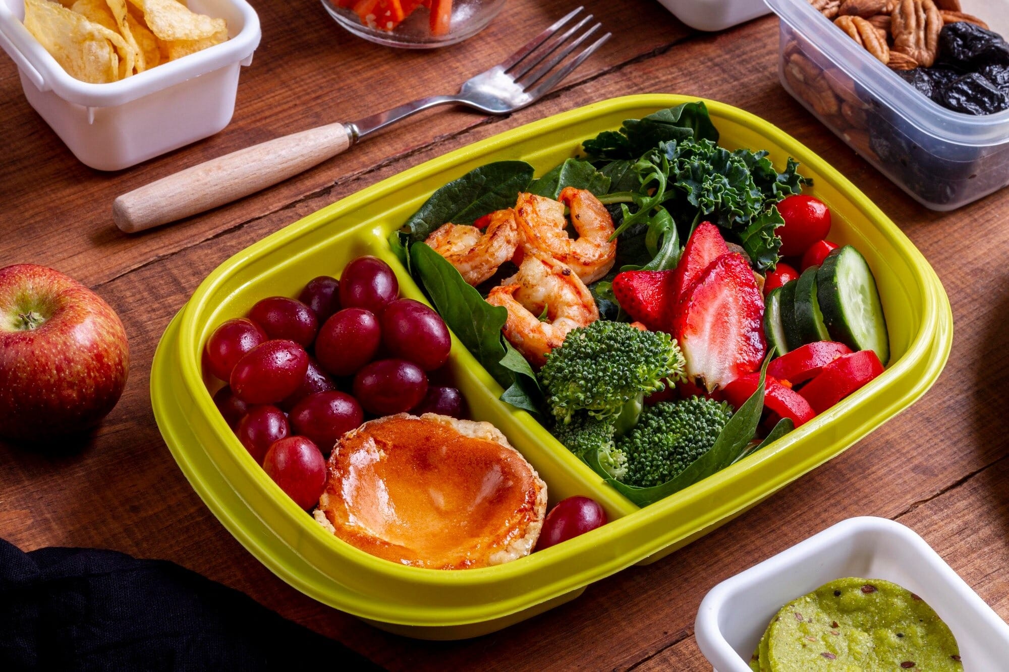 Marmita saudável com camarões grelhados, brócolis, folhas verdes, morangos, pepino, uvas roxas e panqueca pequena, disposta em pote verde sobre mesa de madeira, com lanches e frutas ao redor.