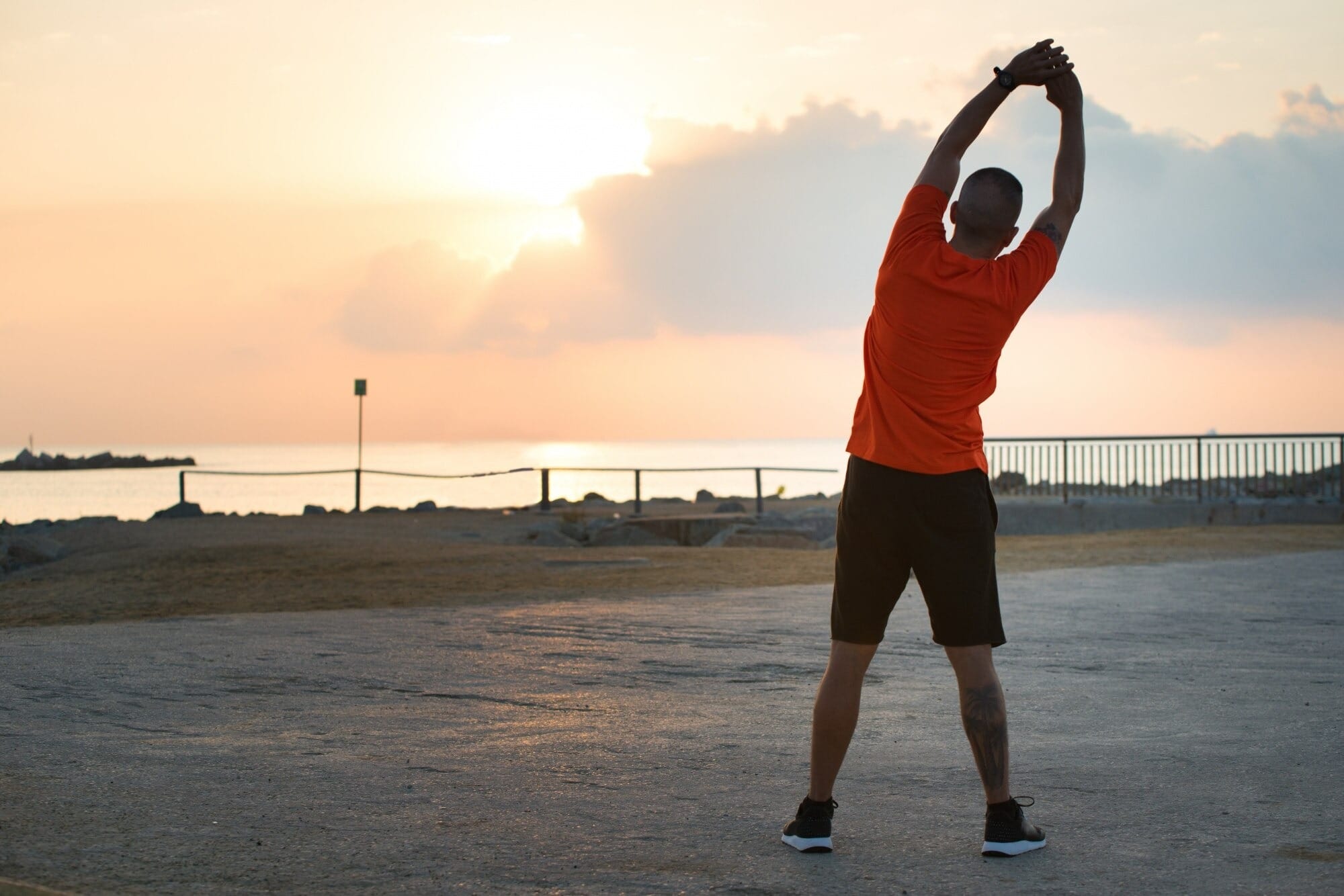 Homem de costas fazendo alongamento com os braços estendidos acima da cabeça em uma orla à beira-mar ao amanhecer, usando camiseta laranja e shorts pretos, com o sol nascendo no horizonte e o mar ao fundo.