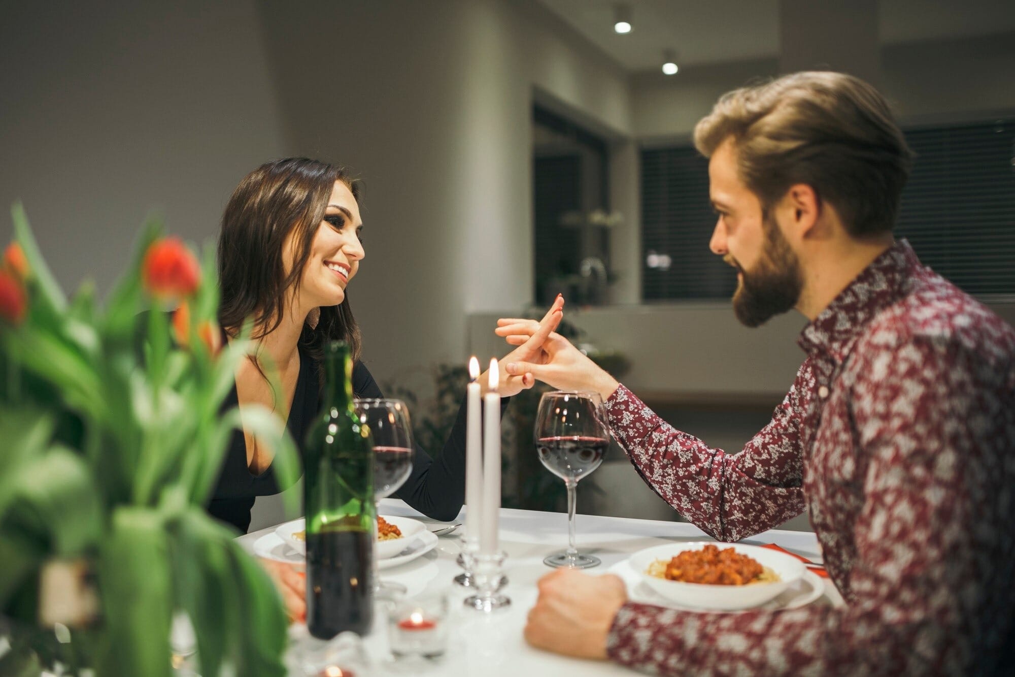 Casal jovem em jantar romântico em casa com velas acesas, taças de vinho tinto e flores na mesa decorada, compartilhando momento íntimo durante refeição especial