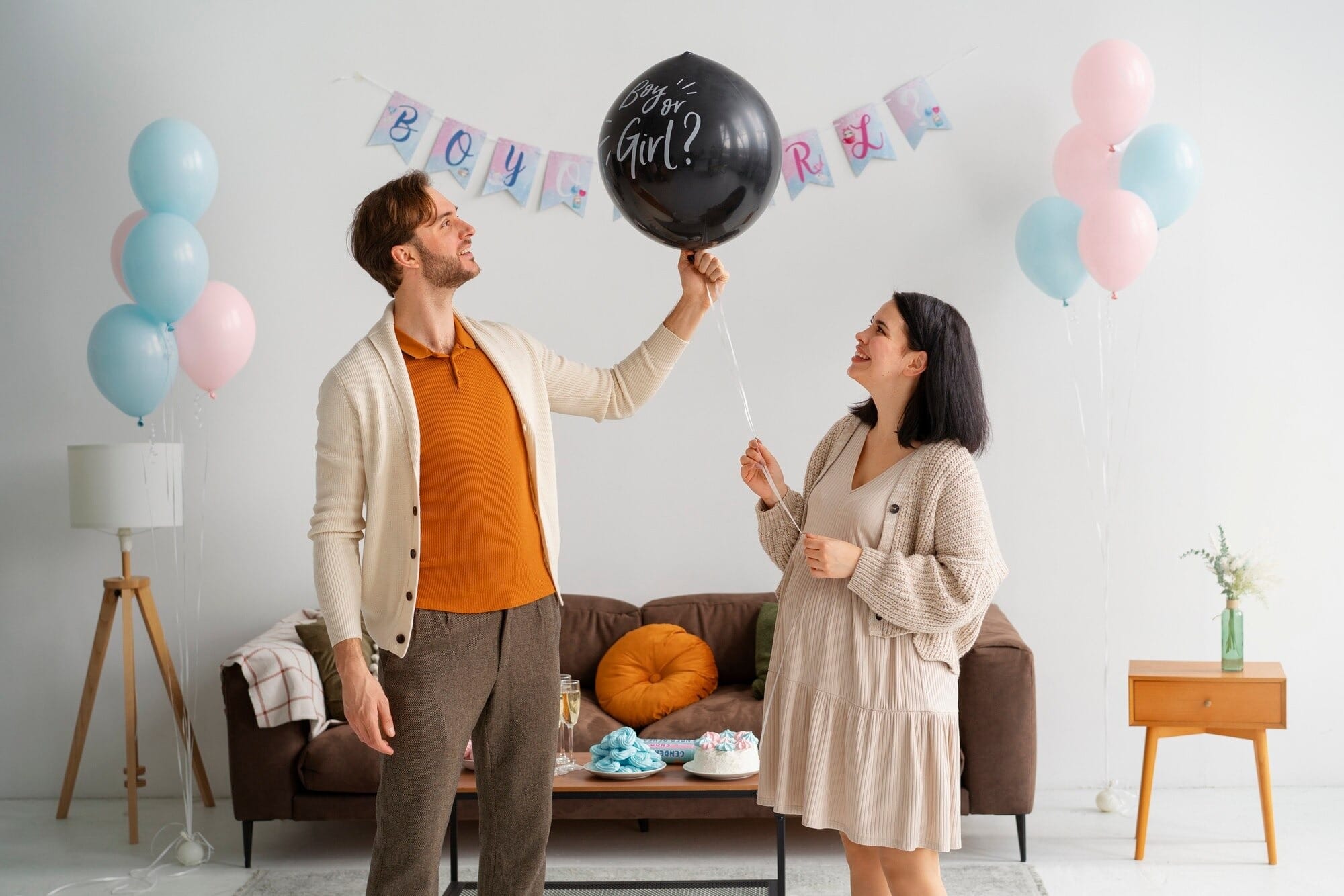Futuros pais em momento de chá de revelação com balão de revelação preto, decoração com balões rosa e azul e bandeirolas Boy or Girl