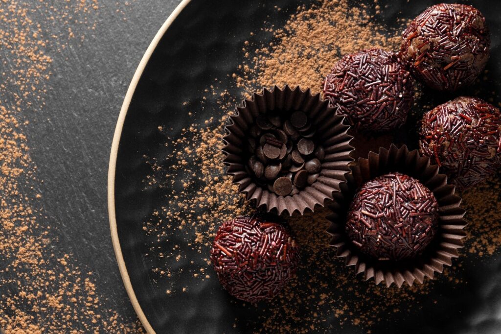 Brigadeiro gourmet de chocolate com granulado e gotas de chocolate servido em forminhas escuras sobre prato preto com cacau em pó