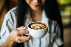 Mulher sorridente segurando uma xícara branca de café com latte art em formato de coração, representando o café perfeito preparado com técnicas de barista