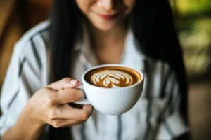 Mulher sorridente segurando uma xícara branca de café com latte art em formato de coração, representando o café perfeito preparado com técnicas de barista