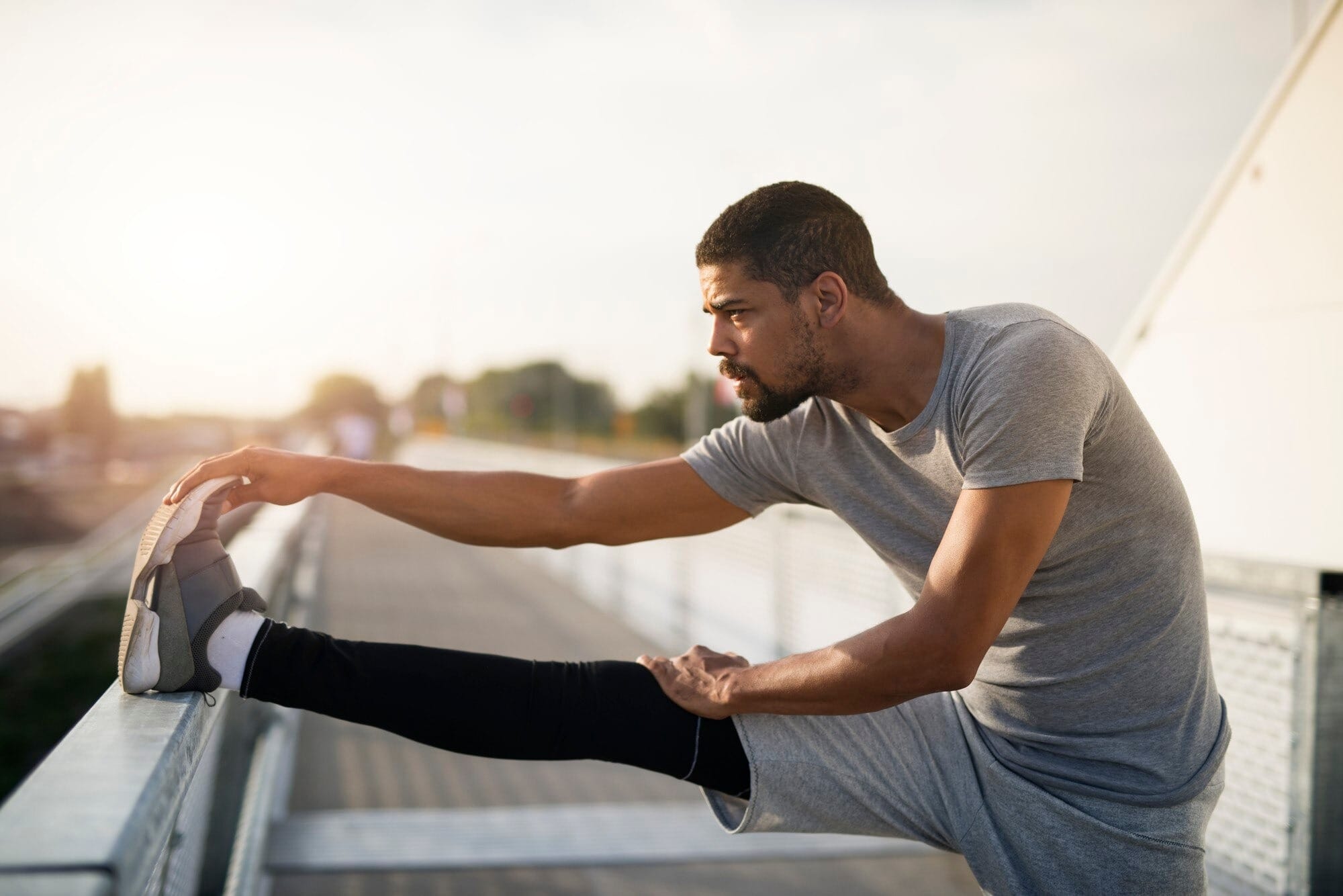 Homem praticando alongamento de flexibilidade para pernas ao ar livre, exercício de isquiotibiais com pé apoiado em superfície elevada