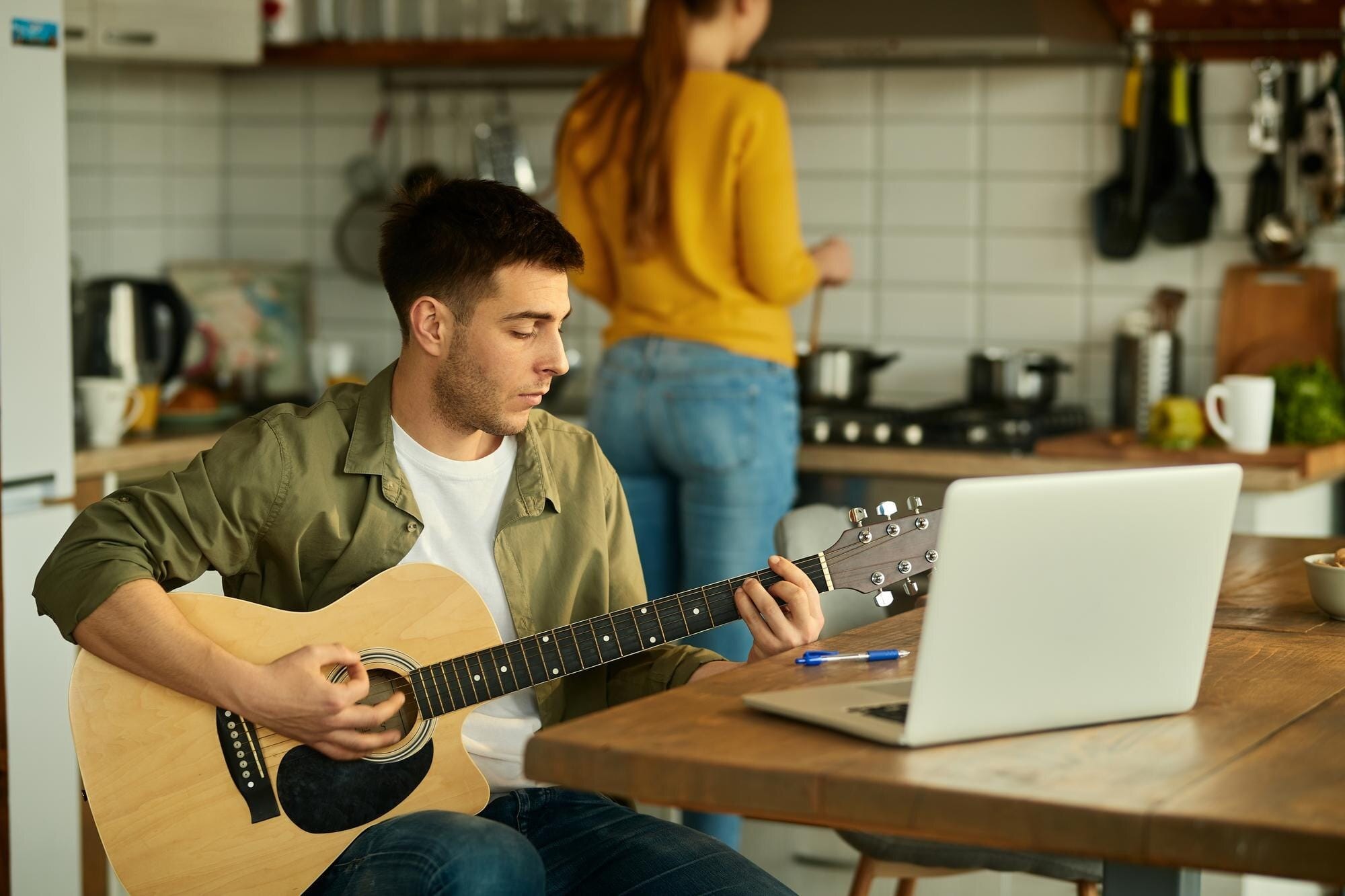 Homem tocando violão acústico em casa enquanto assiste aula online no laptop, representando hobbies criativos e musicais para começar em 2026