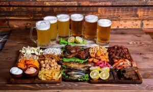 Petiscos para cerveja variados em tábua rústica com batata frita, frutos do mar, carnes, torresmo e cinco copos de cerveja gelada ao fundo