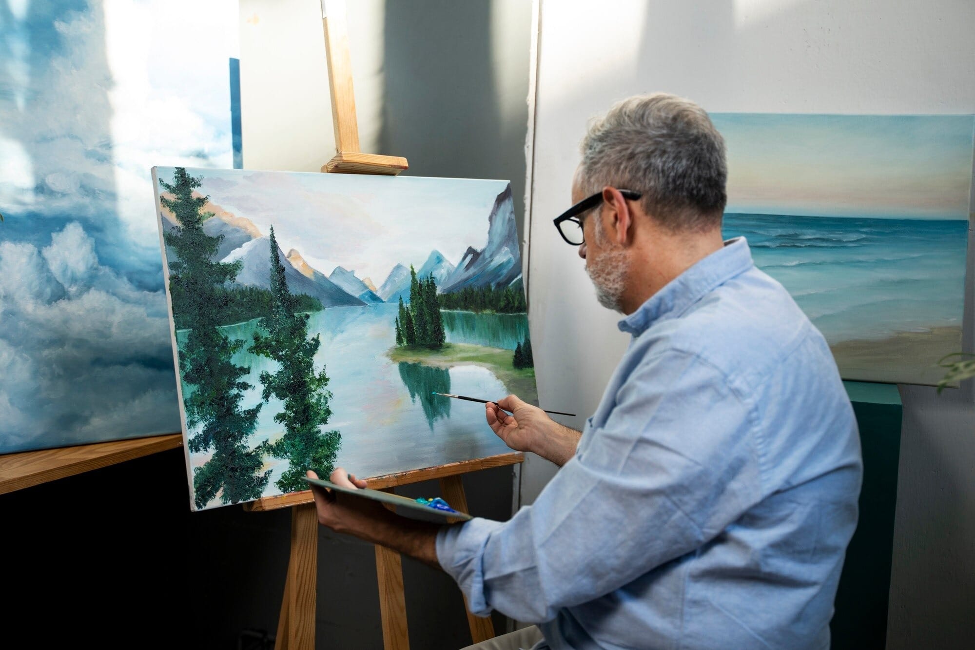 Homem pintando paisagem em tela com montanhas, lago e pinheiros usando técnica de pintura a óleo sobre cavalete em ateliê