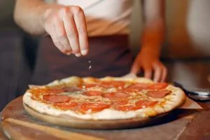 Pizza caseira de calabresa com tomate sobre tábua de madeira, com pessoa temperando com orégano