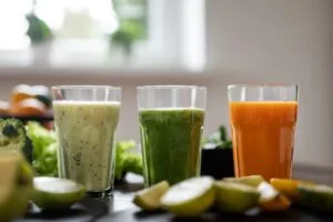 Três sucos detox coloridos em copos de vidro sobre mesa com ingredientes naturais frescos - suco verde, amarelo e laranja para desintoxicar o corpo