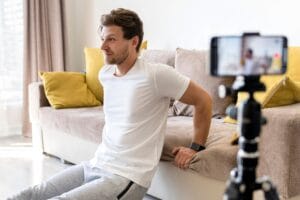 Homem jovem fazendo treino de costas em casa usando o sofá como apoio, enquanto se filma com o celular no tripé na sala de estar.