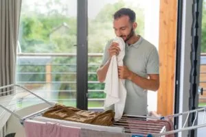 Homem cheirando roupa limpa recém-seca em varal dobrável de chão próximo à sacada de apartamento